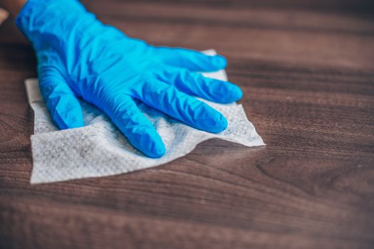 Woman's hand in blue gloves sanitizing cleaning home office wood table surface with wet wipes