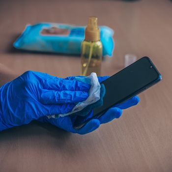 Woman's hand in blue gloves sanitizing cleaning smartphone mobile phone on wood table surface with wet wipes and alcohol
