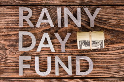 words RAINY DAY FUND laid on wooden surface by metal letters with small roll of us dollar banknotes - flat lay above view