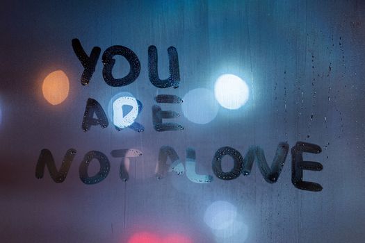 the phrase you are not alone written by finger on night wet glass with blurred classic blue lights in background.
