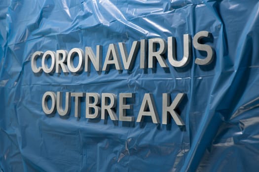 The words coronavirus outbreak laid with silver metal letters on crumpled blue plastic film with perspective and selective focus. Pandemic flu concept.