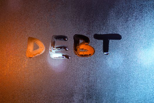 the word debt handwritten on night wet window glass close-up with blurred background.