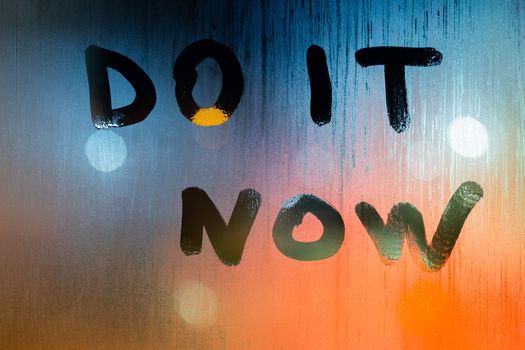 The words do it now handwritten by finger on wet window glass at night with selective focus