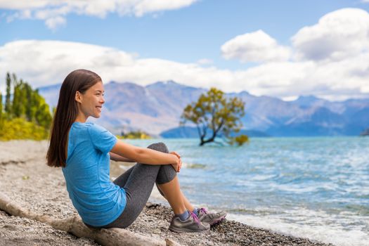 New Zealand Wanaka lake nature landscape travel woman traveler. Asian tourist relaxing enjoying view from beach shore at Wanaka lake landscape with lone tree, famous attraction.