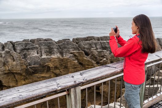 New Zealand travel tourist girl taking smartphone pictures with phone app at Punakaiki Pancake Rocks. Woman in Paparoa National Park, West Coast, South Island, New Zealand