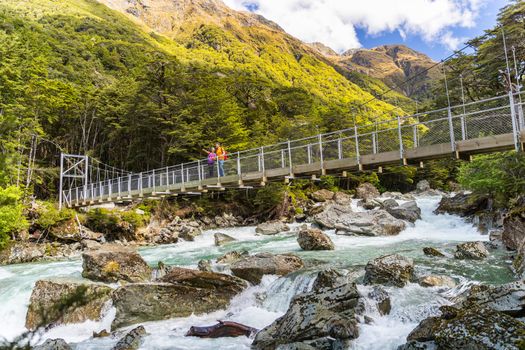 New Zealand hikers tourists crossing river bridge. Couple tramping backpacking woman, man hiking together with backpacks on Routeburn Track trail path.