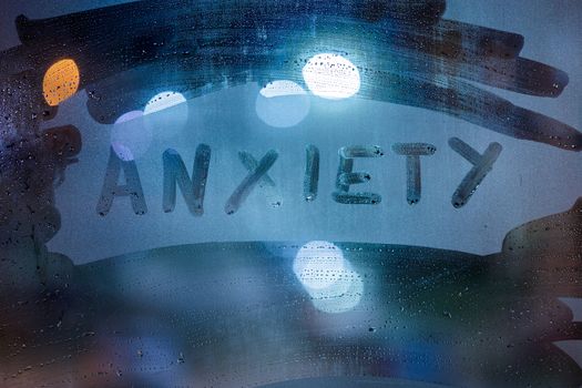 the word anxiety handwritten on night wet glass with classic blue lights in background.