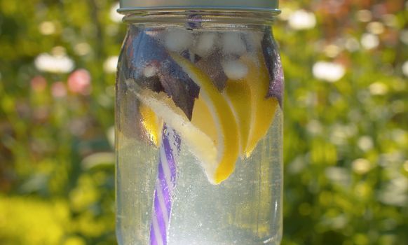 Close up glass misted mug with cold lemonade on blurry natural background outdoors. Macro shooting drink in a country farm at summer day. Healthy lifestyle concept