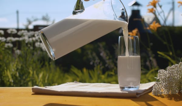 Close up hand pouring milk into a glass. A jug of milk and a glass on a wooden table in the garden of a country house on a background of flowers