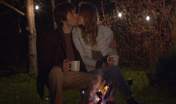 Young happy couple at night sitting near campfire and kissing. Family on weekend in the woods