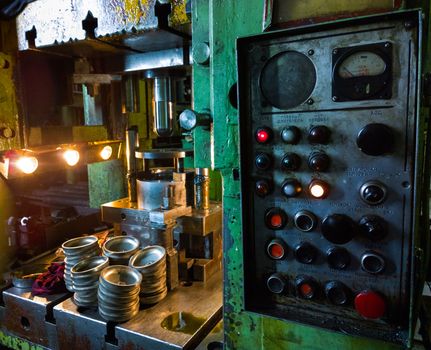 dirty used low tech stamping press producing round aliminium parts in dark environment with large russian control panel.