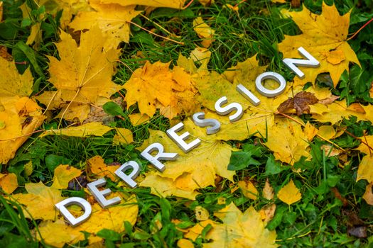 The word DEPRESSION laid with aluminium thick letters on the ground with maple leaves, green grass and diagonal composition. Fall season symbol.