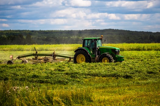 green haymaking tractor on summer field before storm - telephoto shot with selective focus and blur.