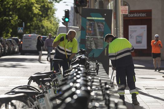 Madrid, Spain - May 19, 2020: Two male workers from the public cleaning and maintenance service are busy cleaning BiciMAD's bicycles in the Retiro district of Madrid.