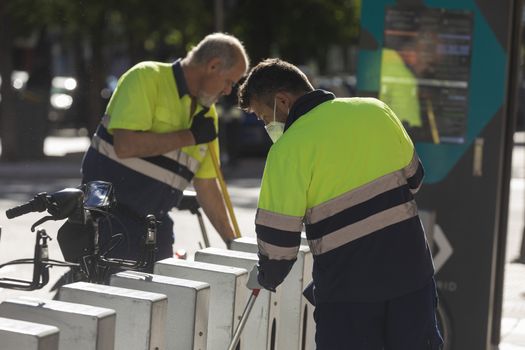 Madrid, Spain - May 19, 2020: Two male workers from the public cleaning and maintenance service are busy cleaning BiciMAD's bicycles in the Retiro district of Madrid.