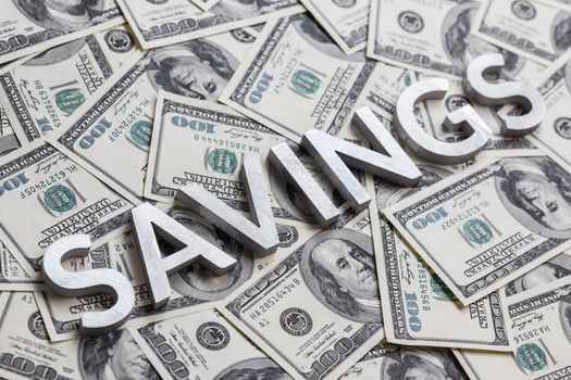 The word SAVINGS laid with white metal letters on the US banknotes background - with selective focus and diagonal composition.