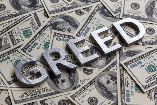 The word GREED laid with aluminium letters on the US dollar banknotes background - with selective focus and diagonal composition.