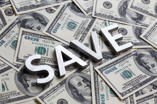 The word SAVE laid with white metal letters on the US banknotes background - with selective focus and diagonal composition.