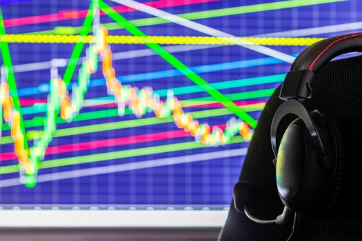 black headset on chair over monitor with trading chart in background - dark background with selective focus and blur