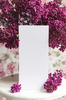 White sheet in blooming purple violet lilac flowers on a white stand on a floral background. Greeting card, place for text, mock up