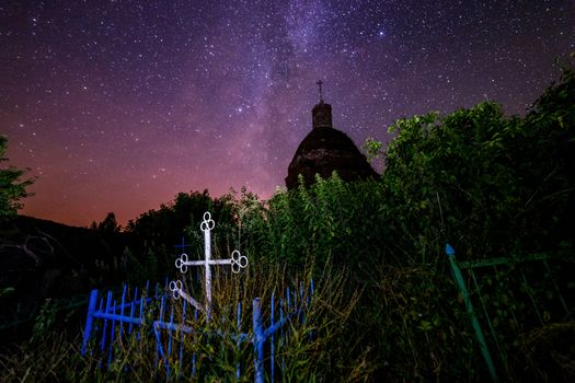 Abandoned christian grave under ruins of church at starry night with visible milky way galaxy. Tall grass and dark bushes selectively focused and lighted. No montage - real photography.