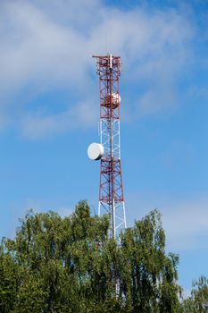 Red-white telecommunication tower with top of green tree on blue sky with clouds background.