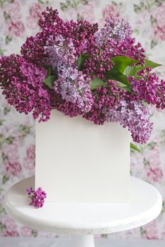 White sheet in blooming purple violet lilac flowers on a white stand on a floral background. Greeting card, place for text, mock up