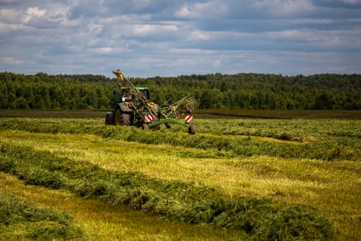 green haymaking tractor on summer field before storm - telephoto shot with selective focus and blur.
