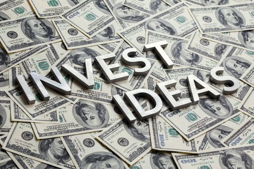 The words INVEST IDEAS laid with thick metal letters on the US dollar bills background - with selective focus and diagonal composition.