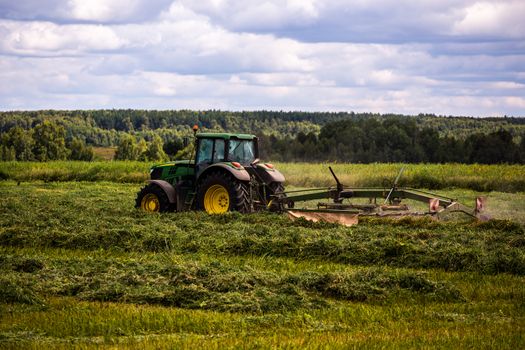 green haymaking tractor on summer field before storm - telephoto shot with selective focus and blur.