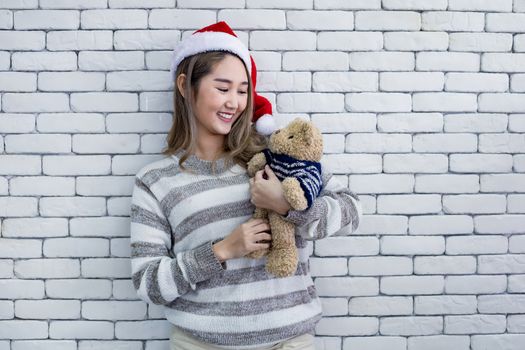 Portrait cute Asian young woman in costume Christmas and wear red santa claus hat and hold a teddy bear with smile on white brick background, copy space. Happy of teen woman in theme Christmas.