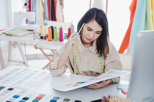 Asian woman designer are thinking and designing clothes for customers order items at the designer desk in the studio. Clothes designers are working in the office. Startup designer concept.