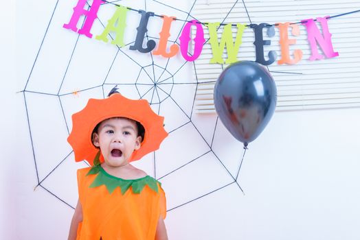 Funny happy little cute boy kid in Halloween costume with orange pumpkin Jack with Cobweb and black balloon in-home on white wall background, Happy Halloween day concept