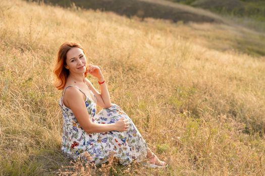 Tender beautiful red-haired girl enjoys the sunset in a field with a hill