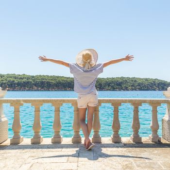 Rear view of happy woman on vacation, wearing straw summer hat ,standing on luxury elegant old stone balcony of coastal villa, relaxing, arms rised to the sun, looking at blue Adriatic sea.