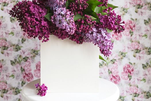 White sheet in blooming purple violet lilac flowers on a white stand on a floral background. Greeting card, place for text, mock up