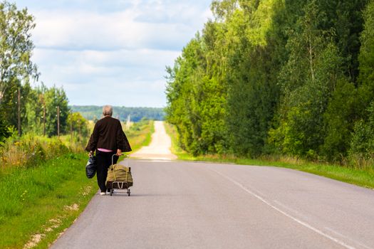old woman with cart and black plastic bag walk away on side of rustic road at cloudy summer day.