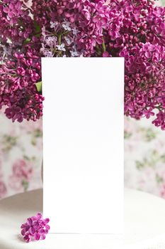 White sheet in blooming purple violet lilac flowers on a white stand on a floral background. Greeting card, place for text, mock up
