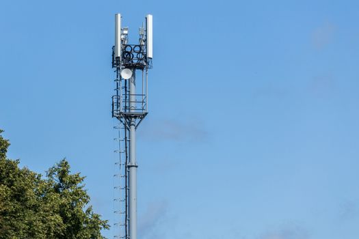 Gray pipe telecommunication tower with top of green tree on blue sky with clouds background.