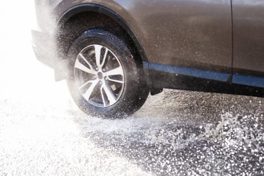 Gray car crossing puddle with water splash at daylight closeup with defocus technique and blur in counter light.