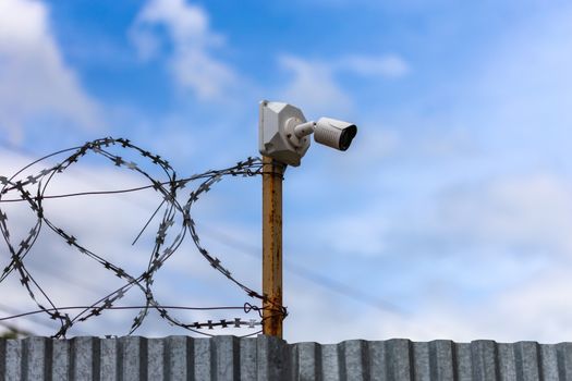 Profiled sheet fence with cheap surveillance camera and barbwire with selective focus at daylight on blue sky with clouds blurred background.