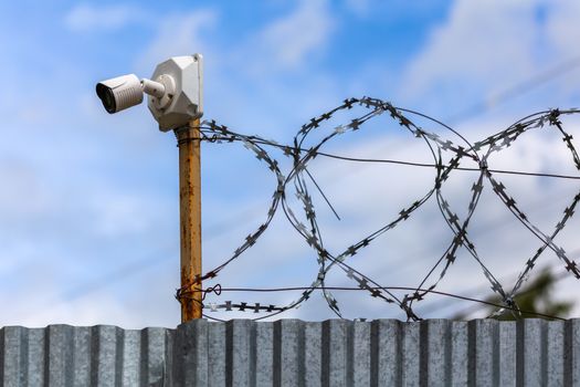 Profiled sheet fence with cheap surveillance camera and barbwire with selective focus at daylight on blue sky with clouds blurred background.