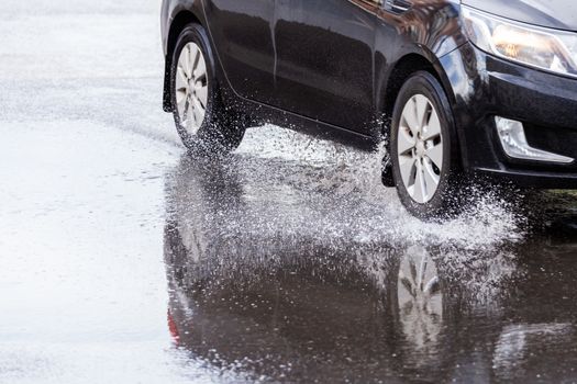 Black car crossing puddle with water splash at daylight closeup with selective focus and blur.