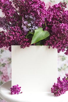 White sheet in blooming purple violet lilac flowers on a white stand on a floral background. Greeting card, place for text, mock up