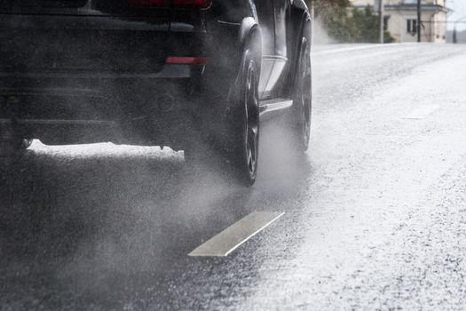 Rain water splash flow from wheels of black car moving fast in daylight city with selective focus. Tiny drops mist.