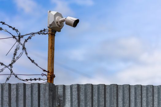 Profiled sheet fence with cheap surveillance camera and barbwire with selective focus at daylight on blue sky with clouds blurred background.