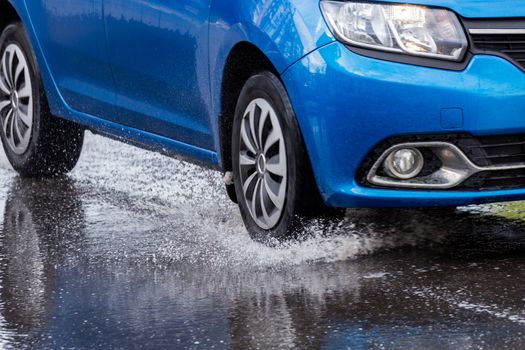 Blue car crossing puddle with water splash at daylight closeup with selective focus and blur.