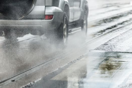 Rain water splash flow from wheels of silver metallic crossover car moving fast in daylight city with selective focus. Tiny drops mist.