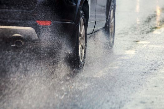 rain water splash flow from wheels of black car moving fast in daylight city with selective focus.