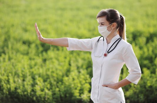 A Female doctor in a medical face mask showing a stop pandemic gesture. Confident female doctor or nurse wearing a face protective mask on green grass background. Coronavirus Covid19. Girl, woman